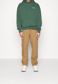 Person som bär grön huva, beige byxor med förstärkt sömnad, vita sneakers, ståendes mot en enkel vit bakgrund.