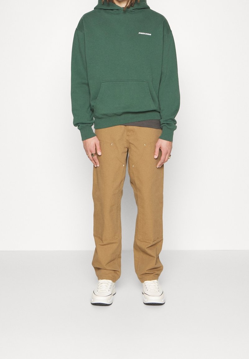 Person som bär grön huva, beige byxor med förstärkt sömnad, vita sneakers, ståendes mot en enkel vit bakgrund.