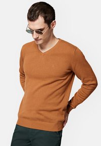 Sweter z dekoltem w serek, z długim rękawem w stonowanym pomarańczowo-brązowym kolorze, z ribbowym wykończeniem. Subtelny akcent w postaci logo na klatce piersiowej. Gładka tekstura.