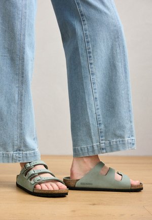 Stopy w jasnozielonych sandałach Birkenstock z korkowymi podeszwami, zestawione z jasnoniebieskimi szerokimi dżinsami na drewnianej podłodze.