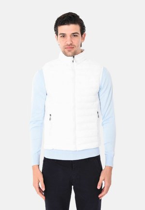 Uomo con camicia azzurra a maniche lunghe, giacca smanicata trapuntata bianca con cerniera e pantaloni neri, in piedi contro uno sfondo chiaro e uniforme.