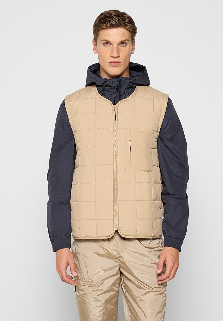 Beige, gestepptes Pufferweste mit Reißverschluss vorne und einer Brusttasche, getragen über einem dunkelblauen Kapuzenhemd, kombiniert mit hellen Hosen.