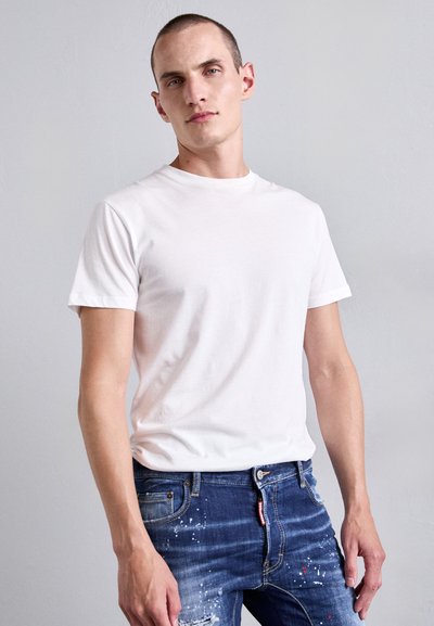 Man with short hair wearing a plain white t-shirt and blue jeans with paint splatters, standing against a light gray background.