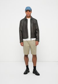 Veste en cuir noire, T-shirt blanc, shorts beiges, casquette bleue, chaussettes noires et chaussures noires ; debout sur un fond blanc.