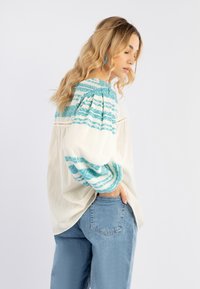 Blusa bianca con accenti turchesi a motivo sulle spalle e sulle maniche, caratterizzata da un taglio morbido e una consistenza delicata, abbinata a jeans azzurri.