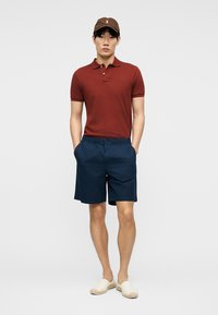 Mladý muž s hnedou čiapkou, tmavomodrým polokošeľou, tmavomodrými kraťasmi a krémovými slip-on topánkami stojí s rukami v kapsách.