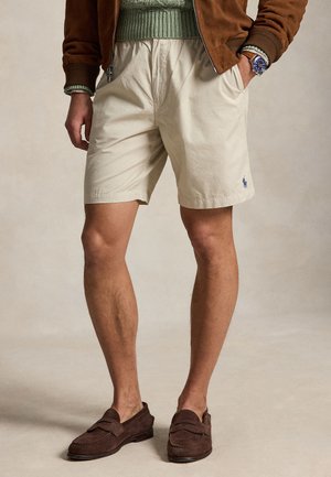 Mężczyzna w beżowych szortach, brązowych zamszowych mokasynach, zielonym swetrze i brązowej kurtce z rękami w kieszeniach na neutralnym tle.