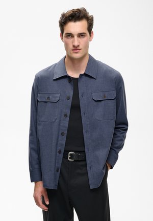 Junger Mann mit braunen Haaren trägt eine dunkelblaue Hemdjacke mit Knöpfen über einem schwarzen Hemd und schwarzen Hosen mit Gürtel, steht vor schlichter weißer Wand.