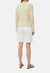 Cremweisser, spitzenstrick Pullover mit gewelltem Saum, kombiniert mit umgeschlagenen weißen Jeansshorts und braunen Riemchensandalen, von hinten betrachtet.