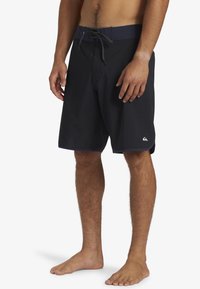 Homme portant un maillot de bain noir jusqu'aux genoux avec une taille à cordon, debout pieds nus contre un fond blanc.