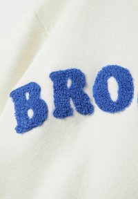Tissu blanc présentant des lettres bleues en relief et texturées formant le mot "BRO" dans un design pelucheux et en relief. Le textile a une apparence douce et une texture uniforme.
