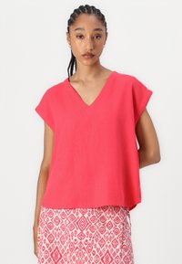 Rotes V-Ausschnitt Oberteil mit kurzen Ärmeln, aus leichtem Stoff, lockere Passform, kombiniert mit einem weißen Rock mit rotem geometrischem Muster.