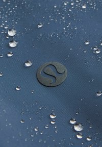 Waterafstotende marineblauwe stof met druppels op het oppervlak, voorzien van een verhoogd zwart cirkelvormig logo in het midden. Gladde textuur met een minimale glans.