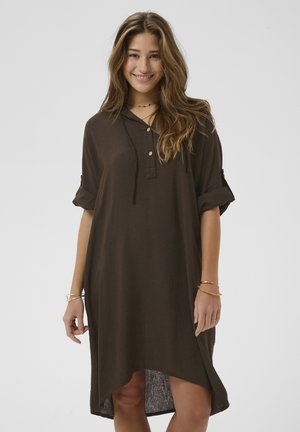 Jonge vrouw die glimlacht, gekleed in een loszittende bruine jurk met opgerolde mouwen en gouden armbanden, staand tegen een effen lichte achtergrond.