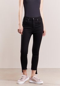 Jeans ajustados negros con una cintura alta, que presentan un dobladillo sin rematar en los tobillos. Combinados con zapatillas de lona rosa claro.