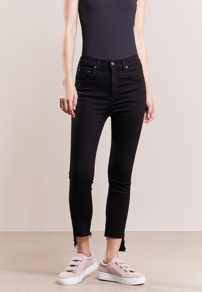 Jeans ajustados negros con una cintura alta, que presentan un dobladillo sin rematar en los tobillos. Combinados con zapatillas de lona rosa claro.