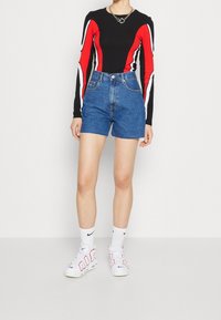 Černý top s dlouhým rukávem s červenými a bílými pruhy, v kombinaci s modrými džínovými šortkami. Modelka má na sobě bílé tenisky a ponožky po kotníky.