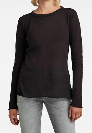 Person iført en ensfarvet sort langærmet trøje og lysegrå jeans, stående mod en hvid baggrund.