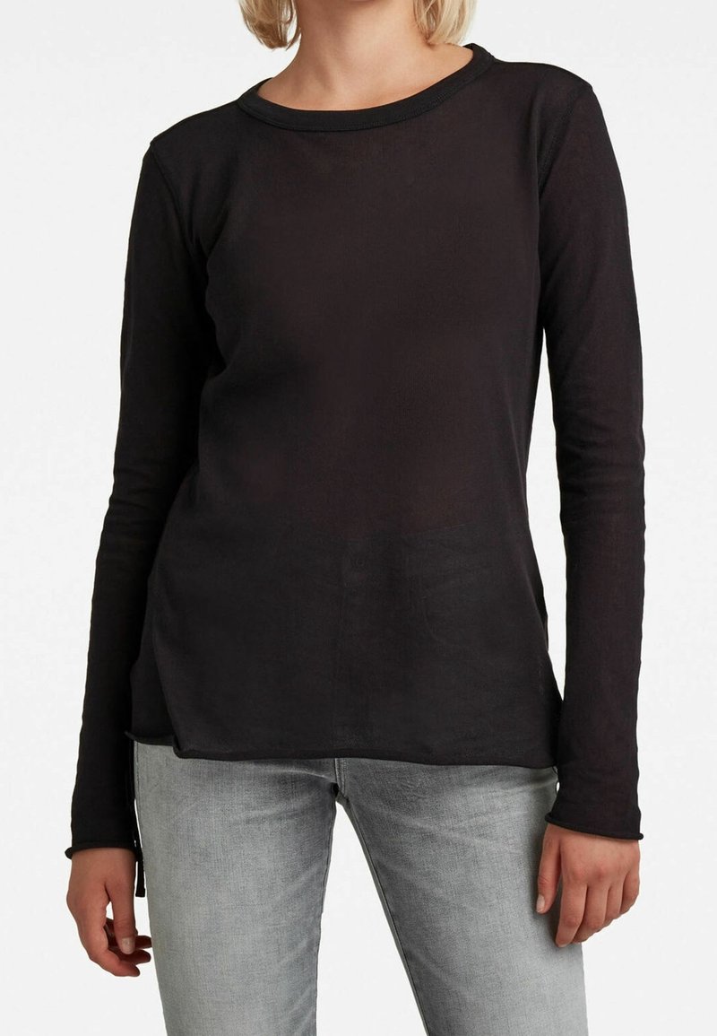 Personne portant un t-shirt noir uni à manches longues et un jean gris clair, debout devant un fond blanc.