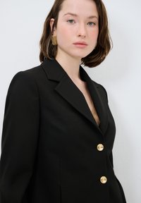 Czarna szyta na miarę marynarka z klapami w kształcie notcha, złotymi guzikami, wyraźnie zarysowanymi ramionami i gładką teksturą materiału. Pod spodem widoczny jest brązowy top.