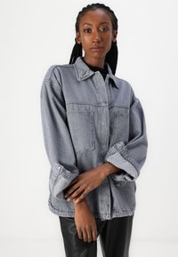 Giacca in denim grigio con colletto, due tasche anteriori e maniche arrotolate. Il tessuto ha una consistenza morbida e una vestibilità comoda.