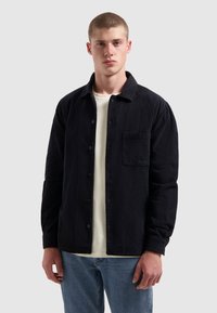Zwarte corduroy shirt met knoopsluiting, lange mouwen en één borstzak, laag over een crèmekleurig T-shirt, gecombineerd met blauwe spijkerbroek.