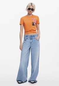 T-shirt in cotone arancione con stampa floreale, abbinato a jeans in denim a gamba larga di colore blu chiaro. Il modello indossa occhiali da sole neri e adotta una postura rilassata.