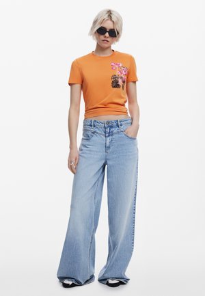 T-shirt in cotone arancione con stampa floreale, abbinato a jeans in denim a gamba larga di colore blu chiaro. Il modello indossa occhiali da sole neri e adotta una postura rilassata.