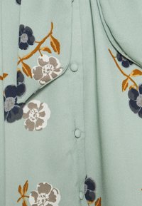 Tissu vert clair avec un motif floral en beige, marine et moutarde, avec des boutons recouverts de tissu et des sections superposées pliées.