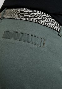 Gröna tygbyxor med en midja i svart och vitt fiskbensmönster samt en bakficka med platt stängning.