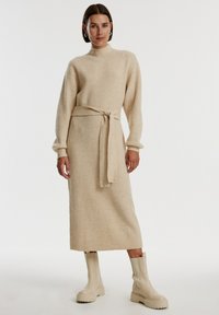 Rippstrickkleid in Beige mit hohem Kragen, langen Ärmeln und gebundenem Taillenbund. Kombiniert mit cremefarbenen Stiefeletten mit dickem Absatz.