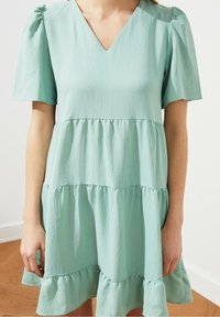 Mátově zelené šaty s vrstveným designem vyrobené z texturované látky, s výstřihem do V, krátkými puff rukávy a volánkovým lemem.