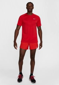 Crvena sportska majica s teksturiranim uzorkom, uparenim s crvenim kratkim hlačama. Crne tenisice za trčanje s crvenim detaljima. Nike logotip vidljiv.