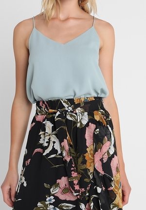 Vrouw draagt een lichtblauw mouwloos topje gecombineerd met een zwarte rok met hoge taille met een meerkleurige bloemenprint en tailleceintuur.