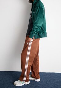 Grüne Velours-Reißverschlussjacke, schwarzes Shirt darunter, braune Hose mit drei weißen Streifen. Die Sneaker haben ein weiß-grünes Design.