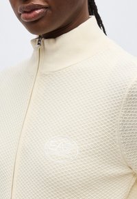 Detailný záber osoby v krémovom textúrovanom pulóvri na zips s vysokým golierom a vyrazeným logom EA7 na hrudi.