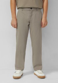 Osoba ubrana w beżowe spodnie straight-leg i białe sneakersy na jednolitym jasnym tle, stoi z rękami w luźnej pozycji.