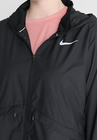 Chaqueta ligera negra con cremallera, con un collar alto y dos bolsillos frontales. Logo de Nike en blanco en el lado izquierdo. Interior suave.