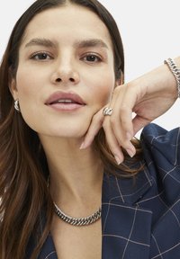 Een zilveren ketting rust om de nek van een model. Ze draagt bijpassende oorbellen en een getailléde donkerblauwe blazer met een ruitpatroon.