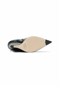 Zapato de tacón alto de piel negra con punta afilada, mostrado desde la suela con fondo beige y marcas de la marca.