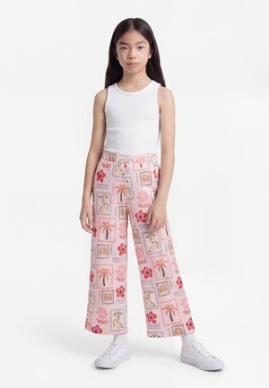 Niña con cabello largo y negro, lleva un top blanco sin mangas, pantalones anchos rosas con estampados tropicales, calcetines blancos y zapatillas blancas.