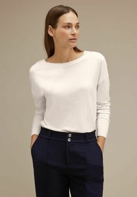 Pullover bianco a maniche lunghe con scollatura ampia, abbinato a pantaloni blu scuro con vita arricciata e due bottoni metallici sul davanti.