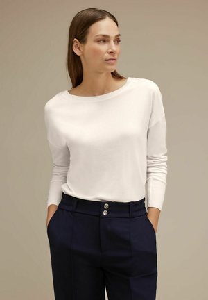 Pullover bianco a maniche lunghe con scollatura ampia, abbinato a pantaloni blu scuro con vita arricciata e due bottoni metallici sul davanti.
