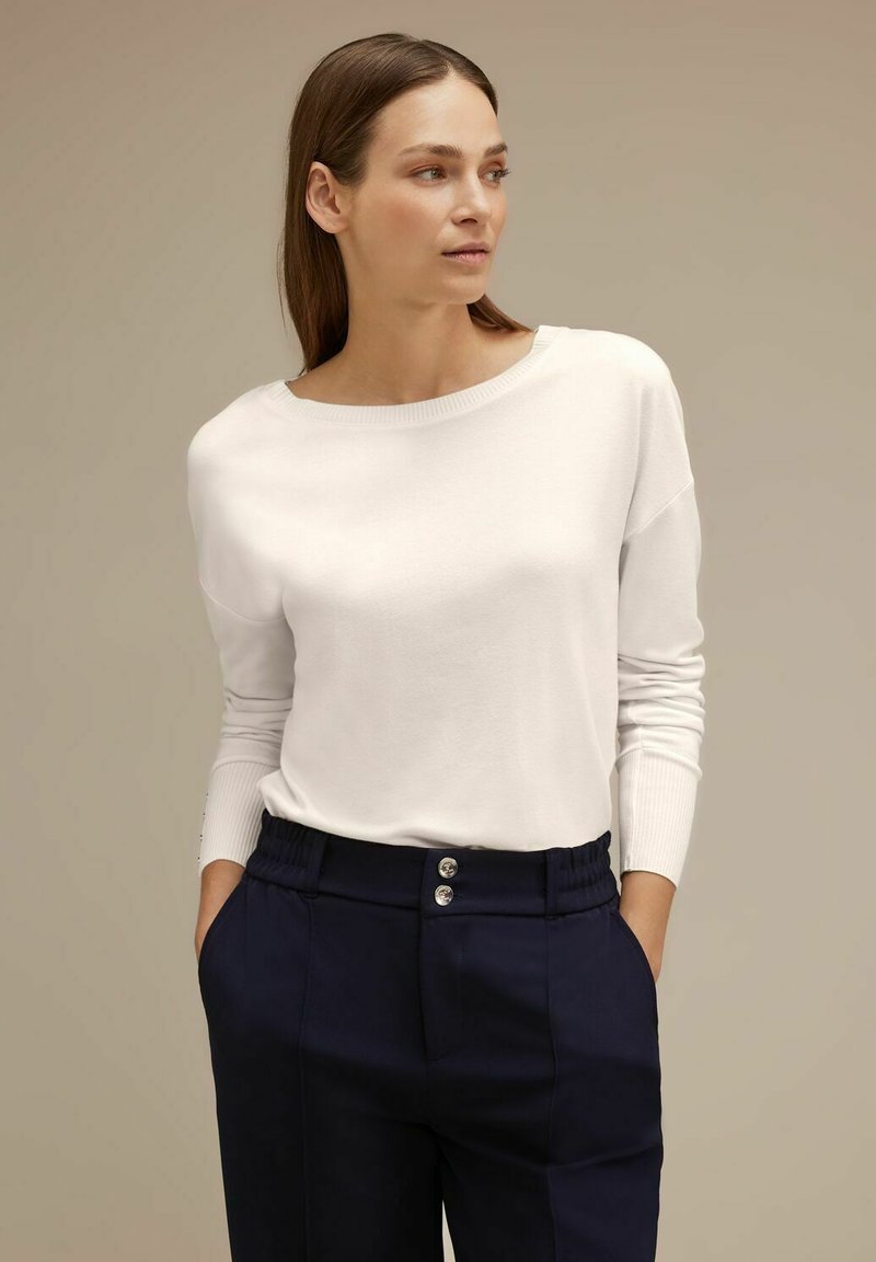 Pullover bianco a maniche lunghe con scollatura ampia, abbinato a pantaloni blu scuro con vita arricciata e due bottoni metallici sul davanti.