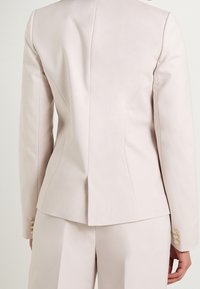 Vaaleanpunainen räätälöity bleiseri, jossa on jämäkät hartiat, sileä pinta ja kultaiset napit kalvosimissa. Takana sauman koristelu.