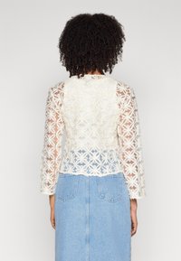 Vit blommig spetsjacka med långa ärmar, som har en genomskinlig textur och rak skärning, i kombination med en ljusblå denimkjol.