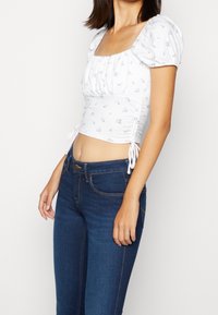 Top corto blanco con estampado floral, mangas abullonadas y fruncido lateral, combinado con vaqueros azul oscuro con costuras en contraste y un corte clásico.