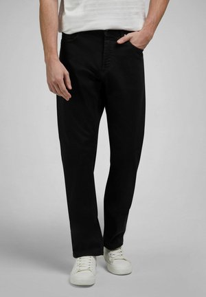 Mężczyzna w czarnych prostych spodniach i białych trampkach, z jedną ręką w kieszeni na jednolitym szarym tle.