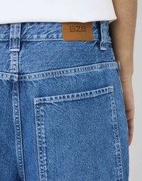 Pantalon en denim bleu avec une texture lisse, présentant une étiquette en cuir marron portant l'inscription "BZD" à l'arrière et deux poches arrière.