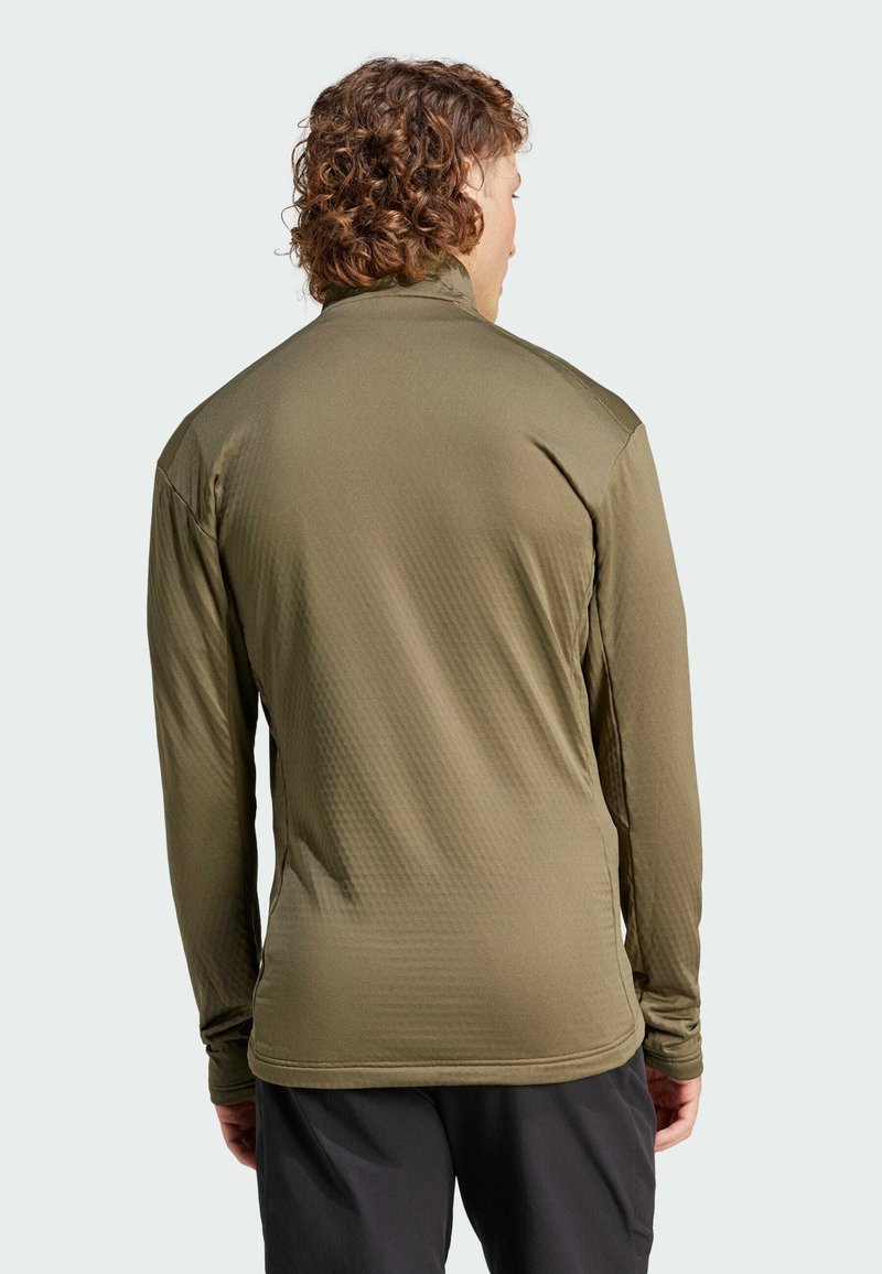 Uomo dai capelli ricci che indossa una maglietta a maniche lunghe verde oliva e pantaloni neri, rivolto di spalle su uno sfondo chiaro e semplice.